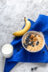 A bowl of oatmeal with blueberries, walnuts, and sliced bananas, next to a glass of milk and a banana on a blue linen napkin.