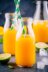 Three glass bottles filled with orange juice, two with green and white striped straws, surrounded by lime slices and ice cubes.