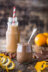 A small glass jar of chocolate pudding with a spoon, next to a bottle of chocolate drink and oranges on a wooden table.