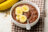 A bowl of chocolate oatmeal topped with sliced bananas and a spoon, with two bananas in the background.
