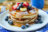 A stack of pancakes topped with strawberries, blueberries, banana slices, and powdered sugar, drizzled with syrup.