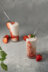 Two tall glasses of strawberry smoothies with white straws and strawberry garnishes, on a gray textured surface.